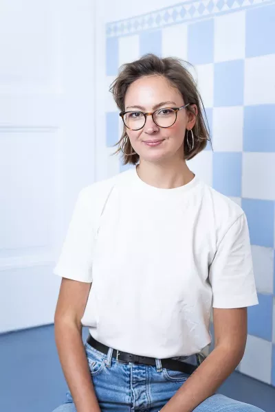 Lea sits in a relaxed position, smiling in the agency in front of a blue and white checkered background with medium-length brown hair, glasses, and earrings, wearing a white T-shirt and blue jeans