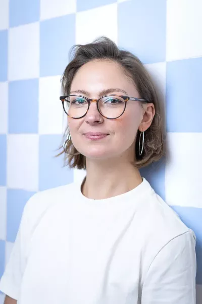 Portrait of Lea with medium-length brown hair, glasses, and earrings wearing a white T-shirt in front of a blue and white checkered background in a bright setting