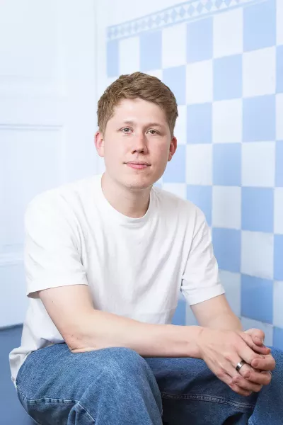 Maxi sits in a relaxed position, smiling in the agency in front of a blue and white checkered background, with short brown hair, wearing a white T-shirt and blue jeans