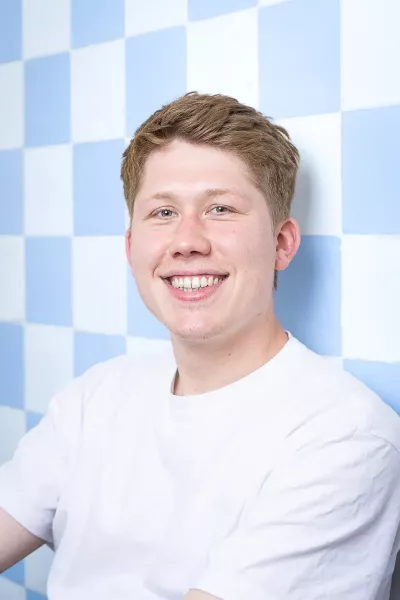 Portrait of Maxi with short brown hair wearing a white T-shirt, smiling in front of a blue and white checkered background in a bright setting