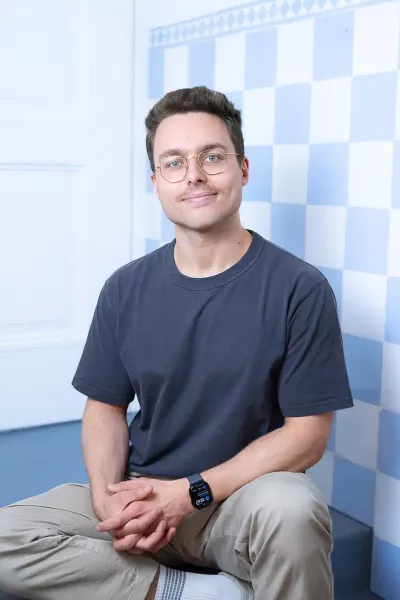 Nico sits in a relaxed position, smiling in the agency in front of a blue and white checkered background, with dark short hair, glasses, and a beard, wearing a dark blue T-shirt