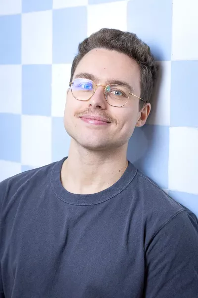 Portrait of Nico with dark short hair, glasses, and beard wearing a black T-shirt, smiling in front of a blue and white checkered background in a bright setting