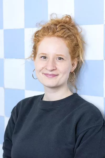 Portrait of Selina with red hair in a bun and earrings, wearing a black sweatshirt, smiling in front of a blue and white checkered background in a bright setting