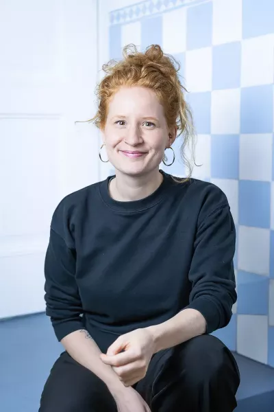 Selina sits in a relaxed position, smiling in the agency in front of a blue and white checkered background, with her red hair in a bun and earrings, wearing a black sweatshirt