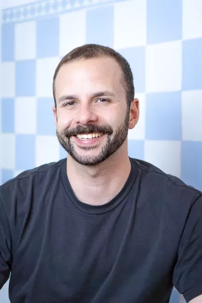 Portrait of Steffen with short dark hair and beard wearing a black T-shirt, smiling in front of a blue and white checkered background in a bright setting