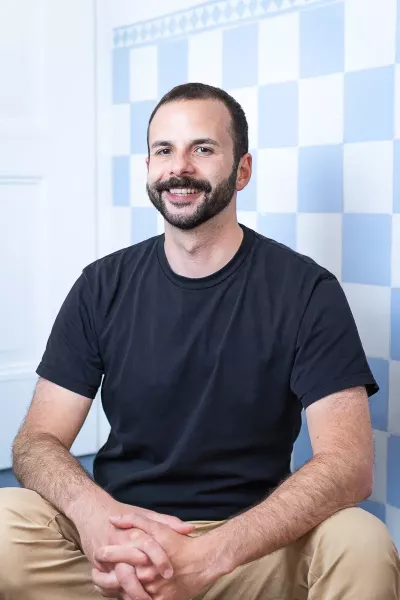 Steffen sits in a relaxed position, smiling in the agency in front of a blue and white checkered background, with short dark hair and a beard, wearing a black T-shirt