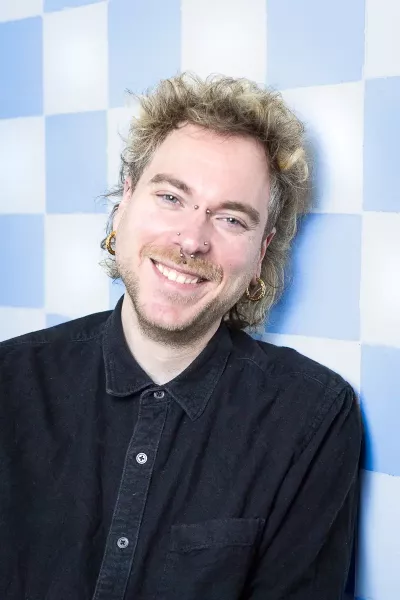 Portrait of Tobi with curly hair, beard, piercings, earrings, and black sweatshirt, smiling in front of a blue and white checkered background in a bright environment