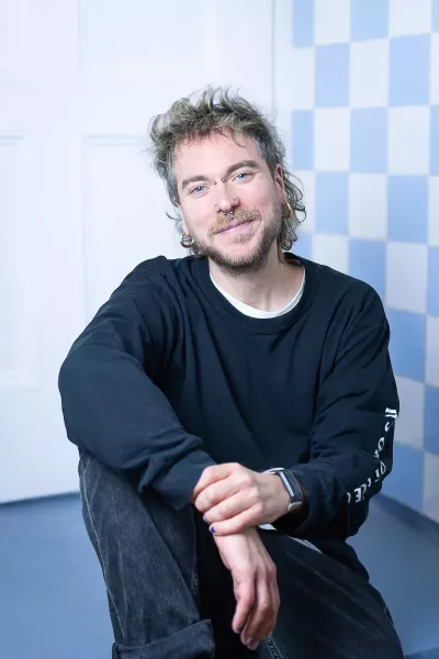 Tobi sits in a relaxed position, smiling in the agency in front of a blue and white checkered background, with curly hair, a beard, piercings, earrings, and a black sweatshirt