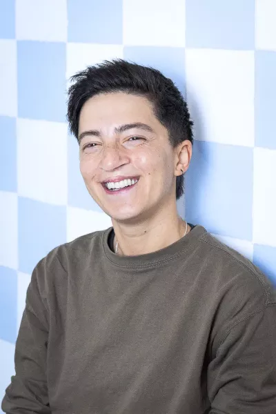 Portrait of Yase with short dark hair and earrings wearing a black T-shirt, smiling in front of a blue and white checkered background in a bright setting