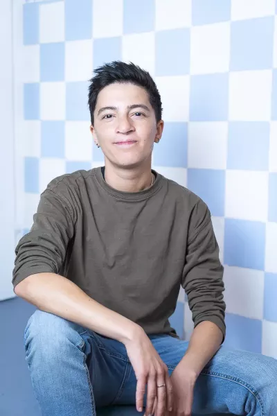 Yase sits in a relaxed position, smiling in the agency in front of a blue and white checkered background, with short dark hair and earrings, wearing an olive green sweatshirt and jeans