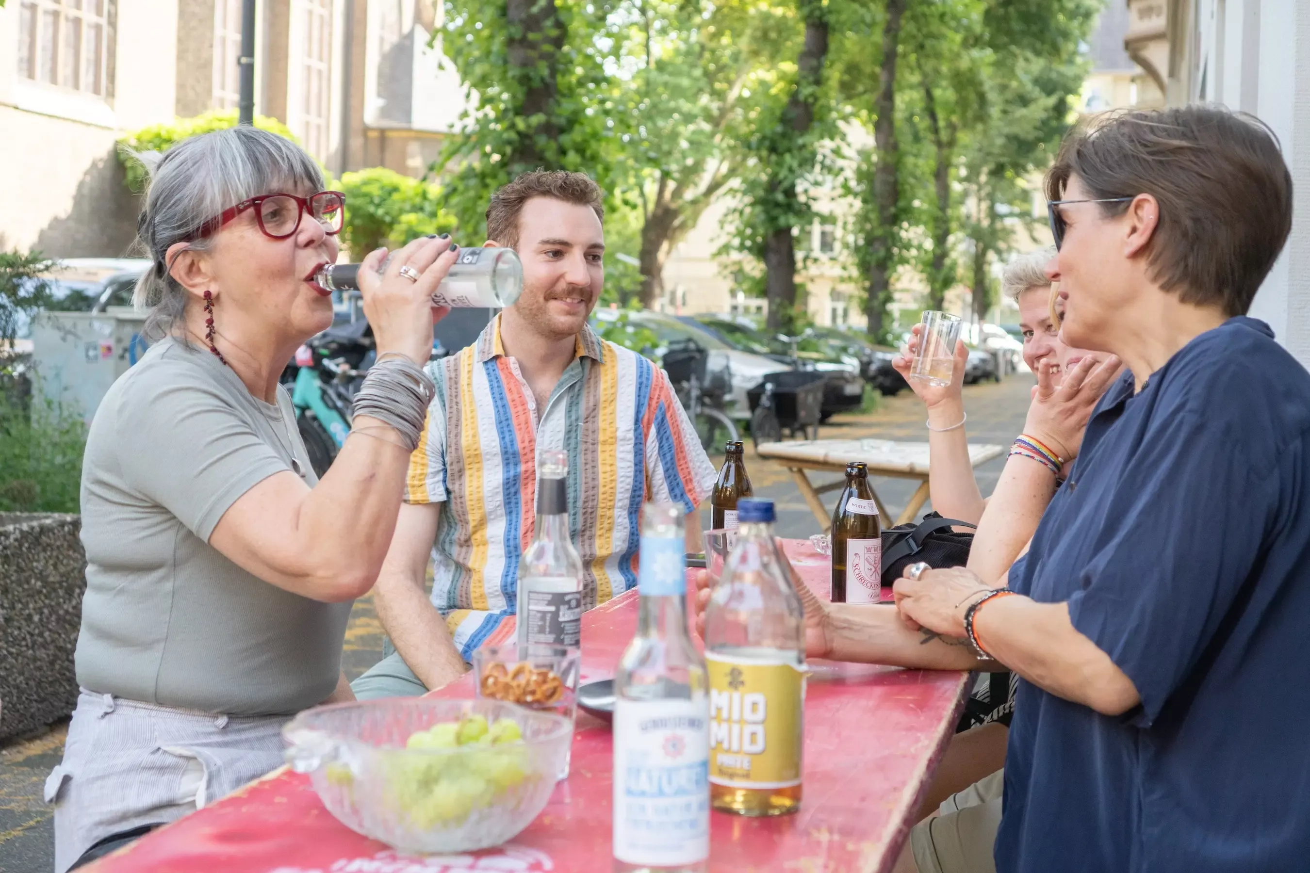 Personengruppe sitzt am roten Tisch draußen, trinkt, lacht und unterhält sich, auf dem Tisch stehen Getränke und Snacks