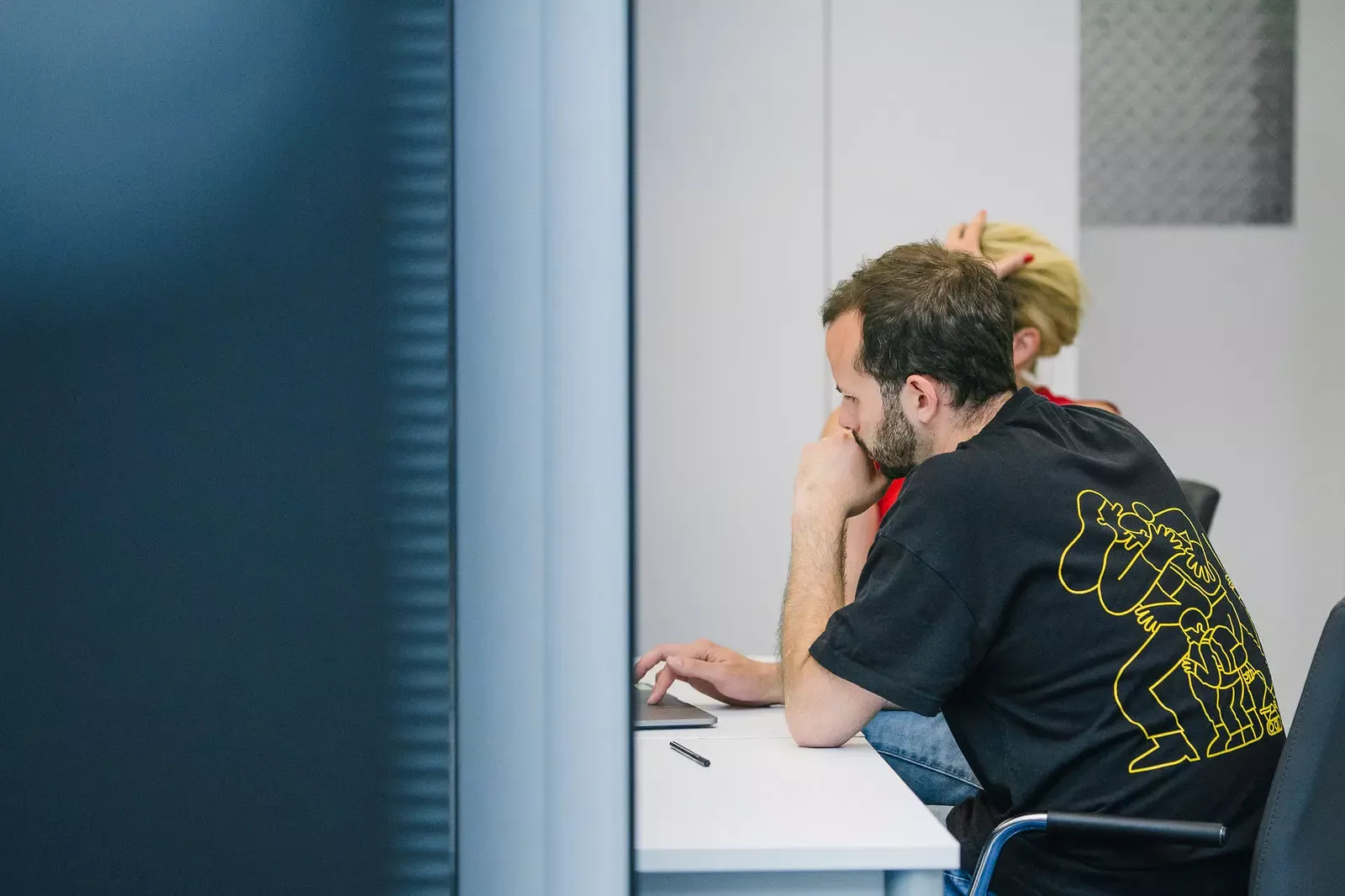 Steffen sitzt konzentriert am Schreibtisch vor Laptop, trägt schwarzes T-Shirt mit gelbem Grafikdruck auf dem Rücken, daneben weitere Person