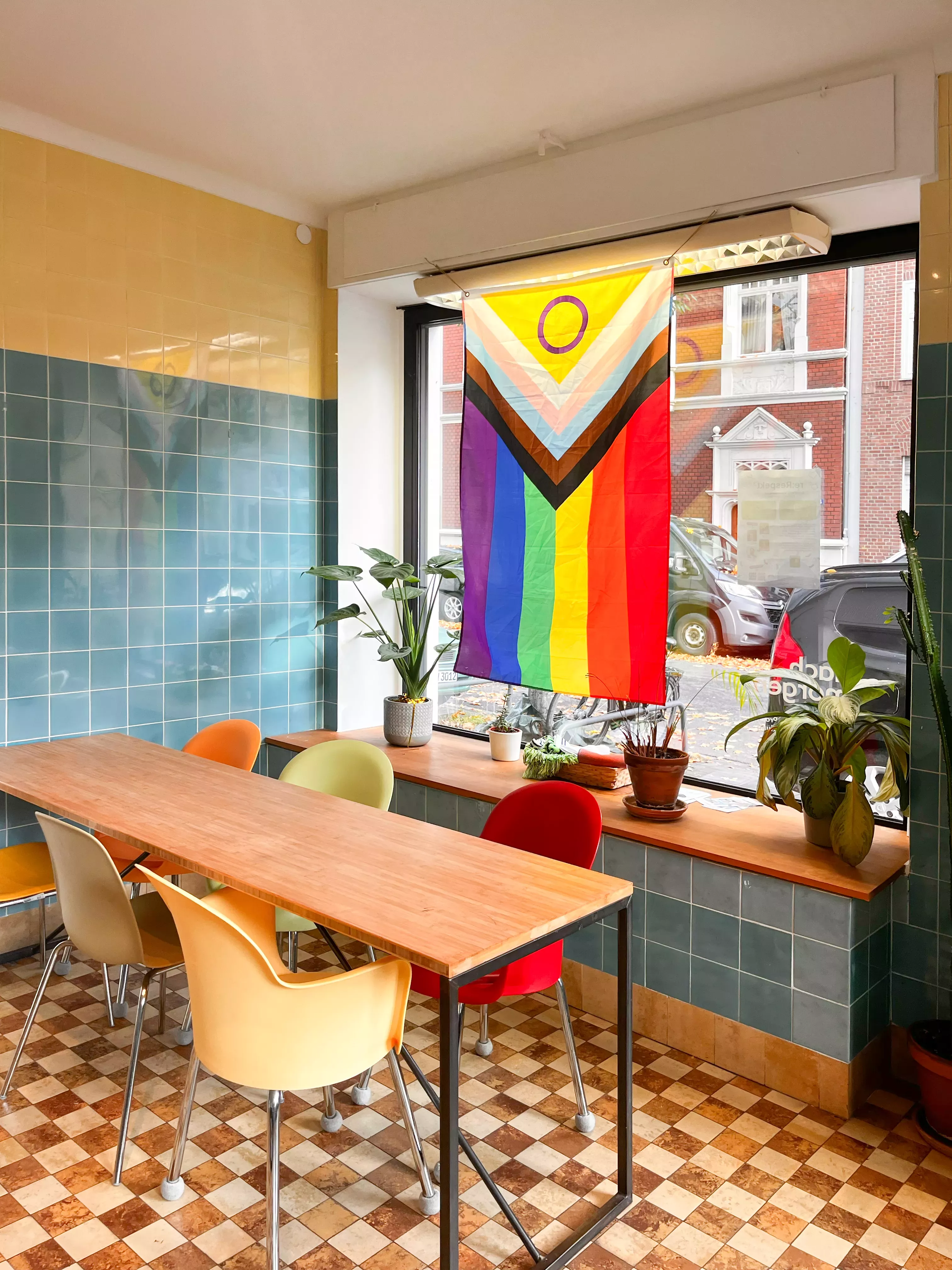 Essbereich im Eingangsbereich vom Büro. Vor dem Schaufenster hängt eine Progressiv-Flag und ein Tisch mit 6 bunten stühlen. Der Raum ist blau, gelb und braun gefließt.