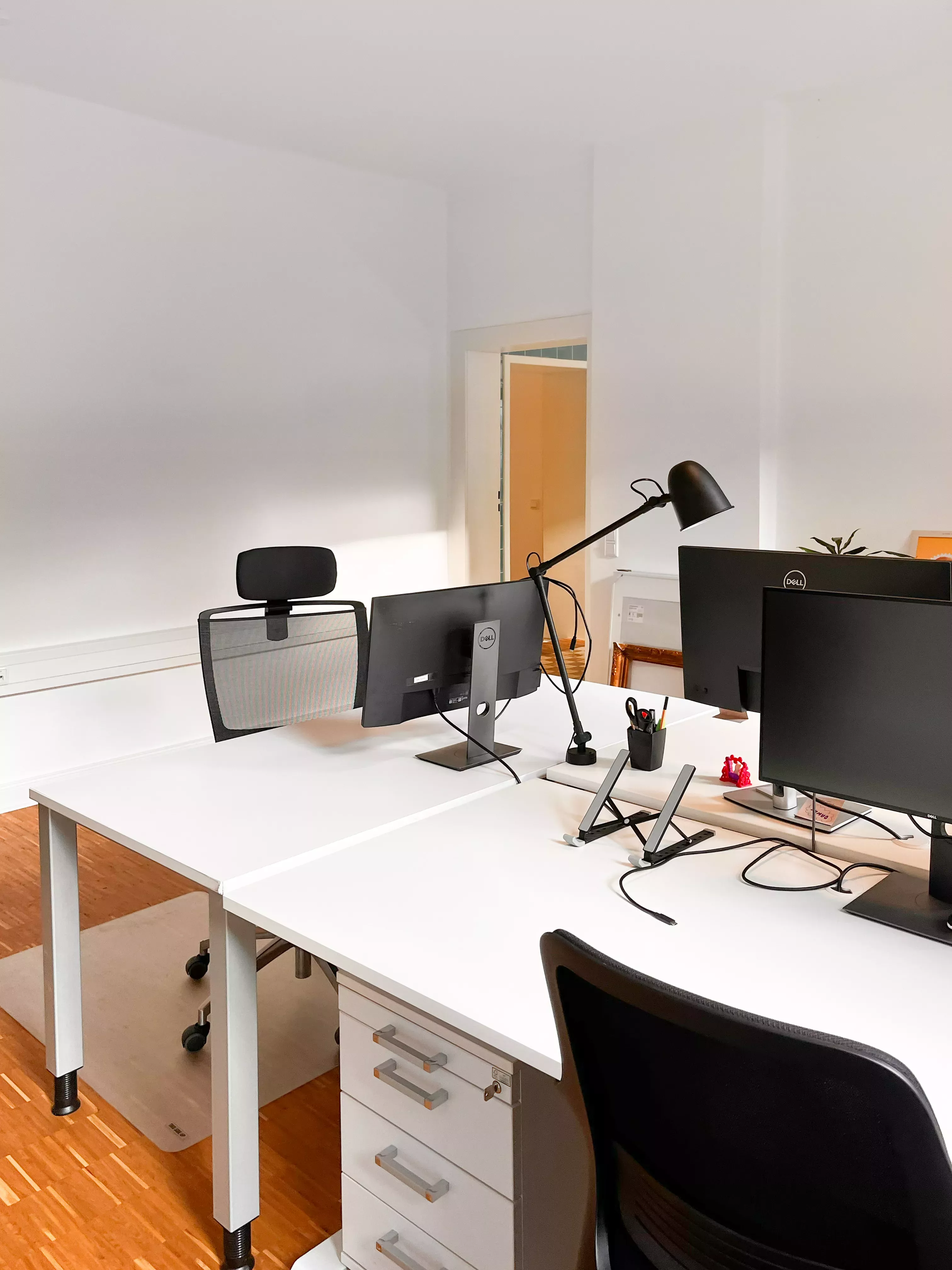 Drei Schreibtische im hellen Büro. Die Tür ist offen und deutet den Eingangsbereich an. Auf den Schreibtischen Stehen Lampen und Bildschirme.