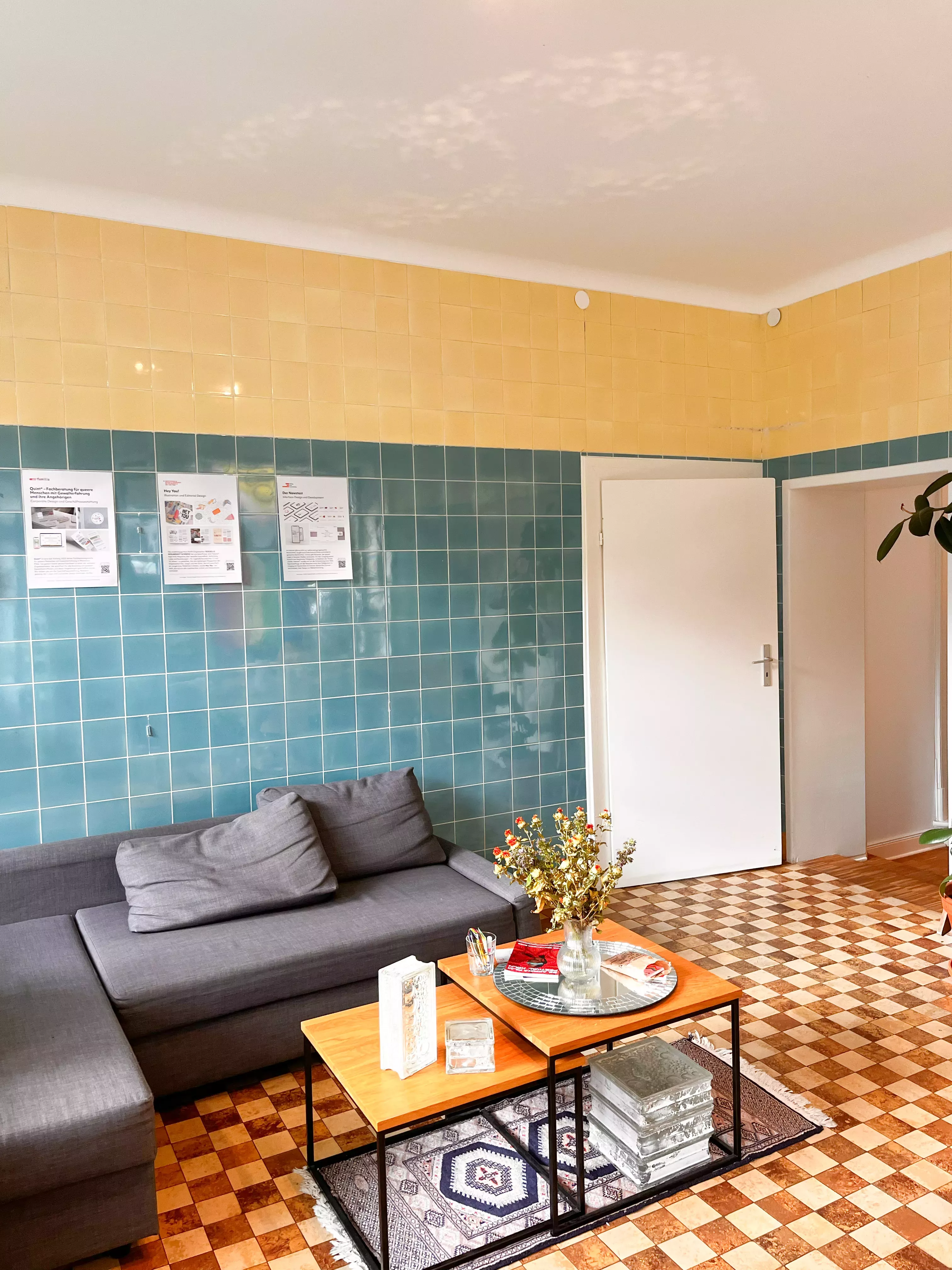 Sitzbereich im Eingangsbereich vom Büro. Es ist ein Sofa mit Beistelltisch abgebildet. Eine Tür führt zu einem Büro und eine Öffnung führt zum Flur. Der Raum ist blau, gelb und braun gefließt.